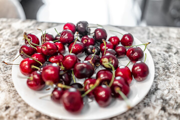 Large fresh sweet cherries washed on white plate with deep red color and green stems closeup with nobody on kitchen island counter table