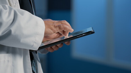 Close up of general practitioner holding tablet computer searching for patient disease report before start planning medication treatment in hospital office. Medical team working night shift