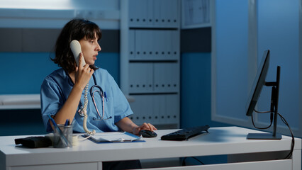 Physician nurse answering landline phone discussing patient report with remote doctor planning medication treatment to cure disease. Medical assistant working over hours in hospital office