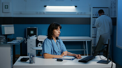 Physician nurse with blue uniform and stethoscope analyzing patient disease report working at health care treatment late at night in hospital office. Assistant checking medical expertise on computer