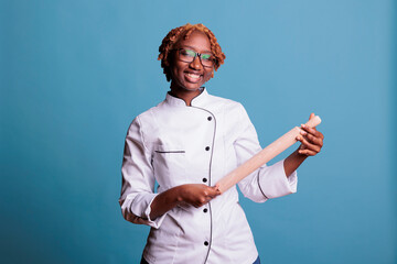 Cheerful african american female baker in uniform and glasses holding dough pin looking at the camera. Joyful cook pretending to play guitar with kitchen utensil in studio shot.