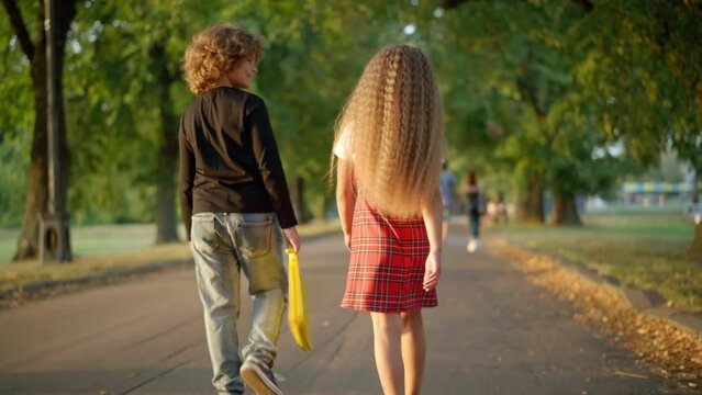 Back view polite Caucasian boy helping girl taking folder walking with friend outdoors. Little gentleman strolling on park alley with sister smiling. Childhood and education