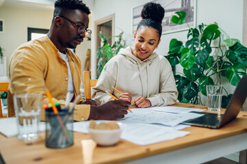 Diverse couple using laptop and looking into the blueprints of their new home