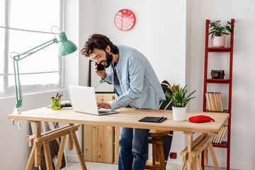 Smiling creative young freelancer professional male talking on mobile phone checking e-mail on laptop computer while working from home office. Business lifestyle concept