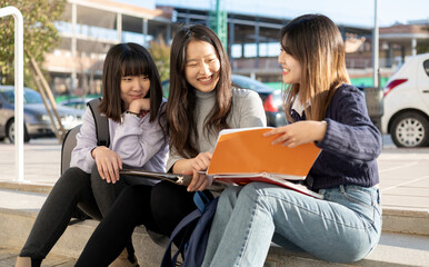 Outdoor student group of Asian girls. The 3 women are sitting on some steps studying, the girl on the right has the book. Education, friendship and people concept. - obrazy, fototapety, plakaty