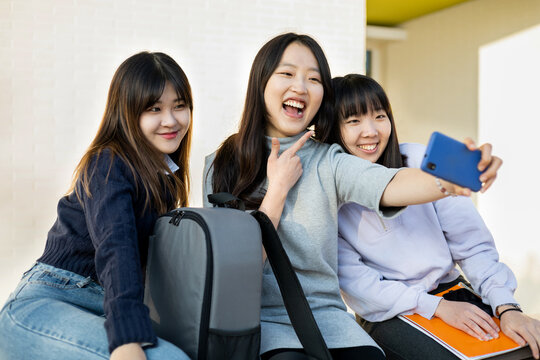 Group of Asian female students outdoors. The 3 women take a photo for social networks, the woman in the middle holds the phone and makes a funny gesture. Education, friendship and people concept.