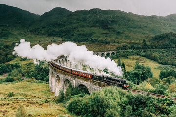 A steam train crossing the Glenfinnan viaduct in the Scottish Highlands made famous by the Harry Potter movies. The Jacobite steam train crossing the bridge with steam in Scotland United Kingdom - obrazy, fototapety, plakaty