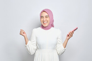 Excited young Asian Muslim woman using mobile phone and celebrating success, getting good news isolated over white background