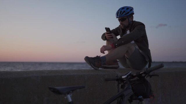 Male cyclist resting on promenade by sea and uses smartphone at sunset. Healthy active lifestyle traveler on bike. Bicyclist stands coast seaview with bicycle build route, looks at map on phone.