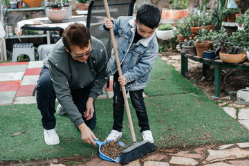 Child helping grandmother and sweeping dry leaves from front yard, using broom and dustpan. Cute boy and senior woman cleaning backyard in springtime.