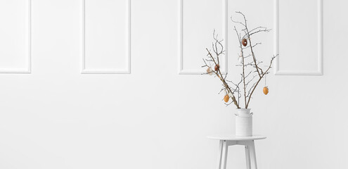 Tree branches with Easter eggs in vase on table near white wall in room