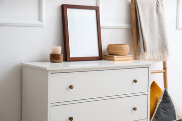 White chest of drawers with blank frame, books and decor near light wall