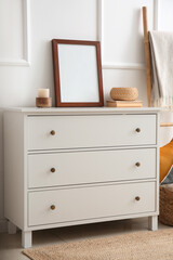 White chest of drawers with blank frame, books and decor near light wall
