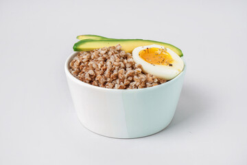 Bowl of tasty buckwheat porridge with soft boiled egg and fresh cucumber isolated on white background