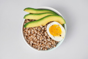 Bowl of tasty buckwheat porridge with soft boiled egg and fresh cucumber isolated on white background
