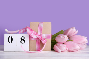 Cube calendar with date MARCH 8, gift box and beautiful tulip flowers on white wooden table near lilac wall. Women's Day celebration