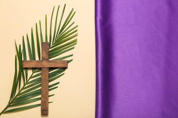 Wooden cross with palm leaf and purple fabric on beige background. Good Friday concept