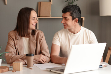 Fototapeta na wymiar Portrait of cheerful young Caucasian woman and man using laptop sitting at table with notebook in modern office, looking at each other, smiling, working together.