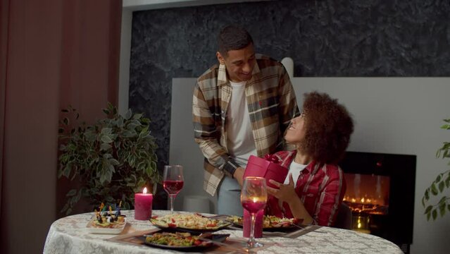 Positive attractive African American man making surprise, giving gift box with present to beloved happy pretty black woman on birthday while cheerful loving couple celebrating event at festive table.