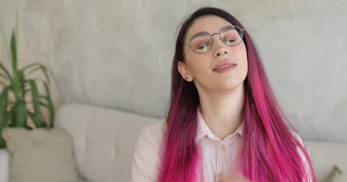 Beautiful smiling caucasian woman with colored long hair standing near the sofa in her living room. She is touching her hair and looking at the camera.