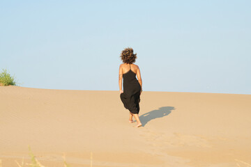 Elegant woman in black dress walking on sand dune. Back view of faceless woman with brunette curly hair in black long dress walking up sand dune against clear blue sky. She is holding her skirt