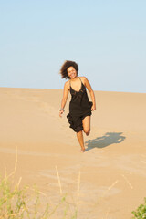 Laughing woman running on sand. Full length portrait of jovial funny woman with dark brown hair and black long dress with bare shoulder, running down the sandy hill laughing out loud.