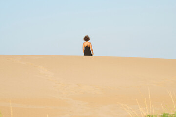 Woman in black walking away in the desert. Back view of anonymous tanned woman in black summer dress and brown frizzy hair walking away from the camera down the sandy hill on blue sky.