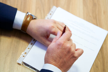 An adult man in an elegant suit holds a ballpoint pen in his hands, studying a paper document....