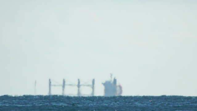 Mirage of cargo ship in the sea beyond the horizon. Ship sailing in the sea. Small waves on the golden surface of warm water with bokeh lights from the sun. Sea, nature and outdoor travel.
