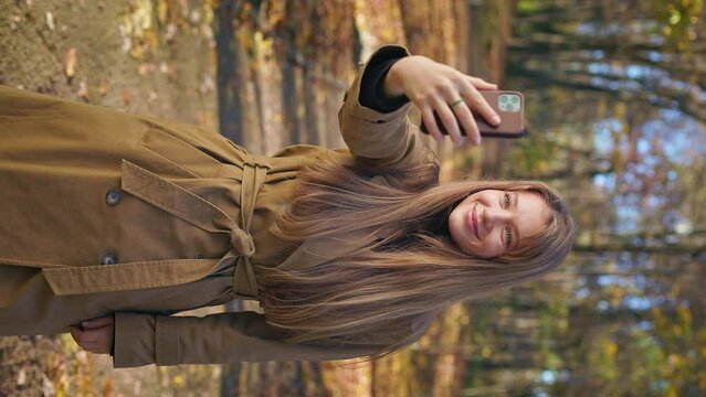 Side view of young, female blogger standing in park, taking selfie video. Attractive lady wearing elegant clothes, smiling, admiring, happy. Concept of modern urban lifestyle.