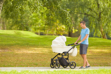 Young adult father pushing white baby stroller and slowly walking on sidewalk at town park in warm...