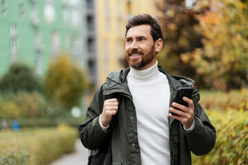 Handsome man with a beard on the city street, holding a phone in his hand. He has a backpack on his...