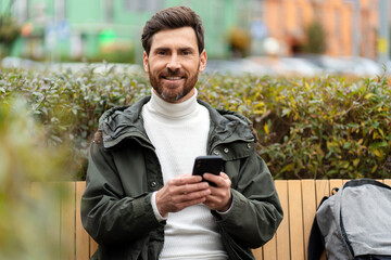 Fototapeta premium Bearded man holds a phone in his hands, looks at the camera, smiles sweetly. A man outside, sitting on a bench.