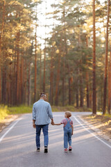 Fototapeta premium Dad and his little daughter are walking along a forest road among tall pines. Family walk in the forest at sunset, man and little girl.