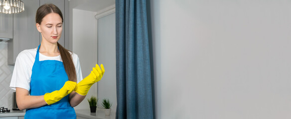 Young woman in uniform puts on yellow rubber gloves to clean the room, copy space. Cleaning concept. Banner