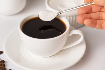 The process of pouring white sugar from a spoon into a white cup of coffee. Sugar addict, diabetes.