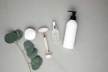 White bottles with facial cosmetics, pink face roller with a branch of eucalyptus on the gray concrete background. Top view