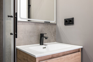 Closeup of a white square ceramic sink on a wooden vanity. Fittings and faucet made of ferrous metal. On the wall is a designer mirror with a white frame. Walls in gray pleasant tones.