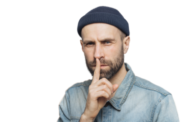 Serious pleasant looking bearded male with mysterious expression keeps fore finger on lips, asks to keep personal information confidential, wears stylish hat and denim shirt, isolated on grey wall