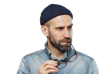 Portrait of pensive bearded man takes off glasses, wears denim stylish jacket and hat, isolated over pink background. Middle aged unshaven male contemplates about something, looks concentrated