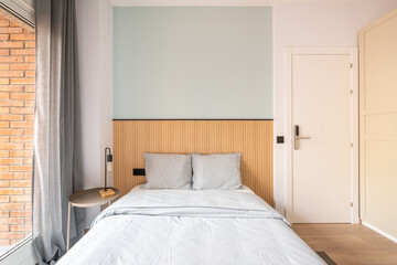 Bed with headboard against wall with wooden decor. Bed is linen in pastel gray. Large window with dark gray curtains. Bedside table with night lamp. Door is locked with an electronic lock without key.
