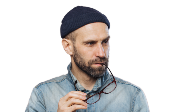 Portrait of pensive bearded man takes off glasses, wears denim stylish jacket and hat, isolated over pink background. Middle aged unshaven male contemplates about something, looks concentrated