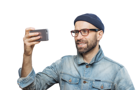 Pleased smiling bearded male with happy expression poses at camera of smart phone, takes photo of himself, wears denim jacket and glasses, isolated over white background. People and technology concept