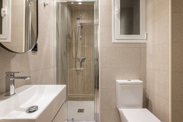 Beautiful fresh and comfortable bathroom for water procedures, morning and evening exercise. Room in beige tones with large sink, beautiful mirror, glass shower and white toilet.