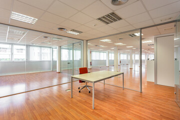 In a bankrupt office building, one of the many empty rooms with transparent, solid glass wall panels and an outdated ceiling covering is in need of repair and refurbishment.
