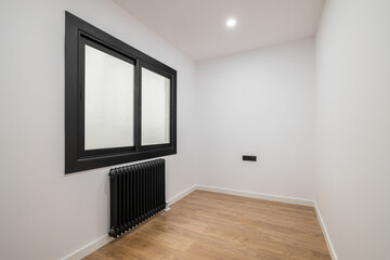 An empty room with a modern version of a plastic window with a black frame and opaque glass. Radiator for heating black. Bright lighting from the ceiling and wooden parquet.