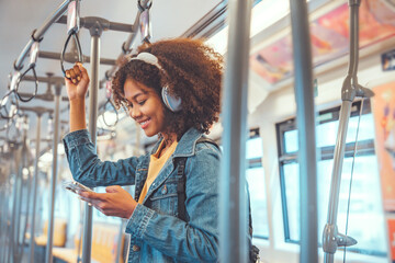 Happy young African American woman passenger listening music via smart mobile phone in a train,...