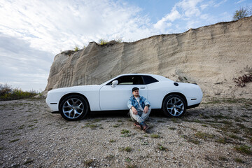 Handsome man in jeans jacket and cap sitting near his white muscle car in career.