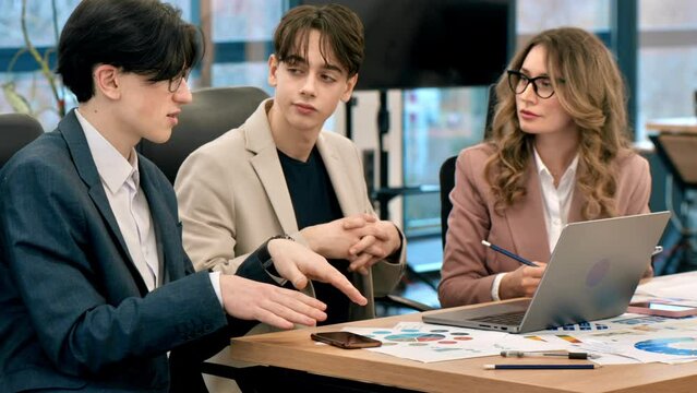 Business meeting in an office, female team leader and two young workers discussing business affairs using laptop. Papers with charts on the table