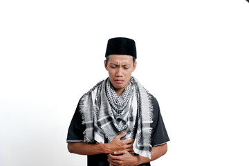 Religious muslim asian man wearing turban, muslim dress and cap, feeling pain due to stomach ache or ulcer. Isolated on white background.